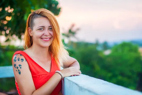 Beautiful red-haired girl in a red dress and tattooed laughs on the veranda of a summer restaurant