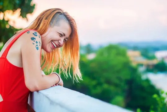 Beautiful red-haired girl in a red dress and tattooed laughs on the veranda of a summer restaurant